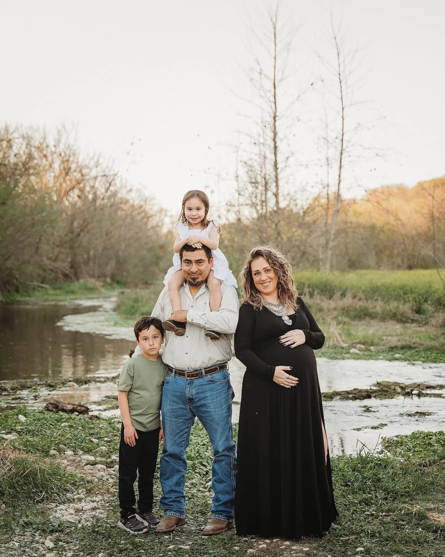 Family posing outdoors: father with a child on his shoulders, pregnant mother, and another child; near a stream.