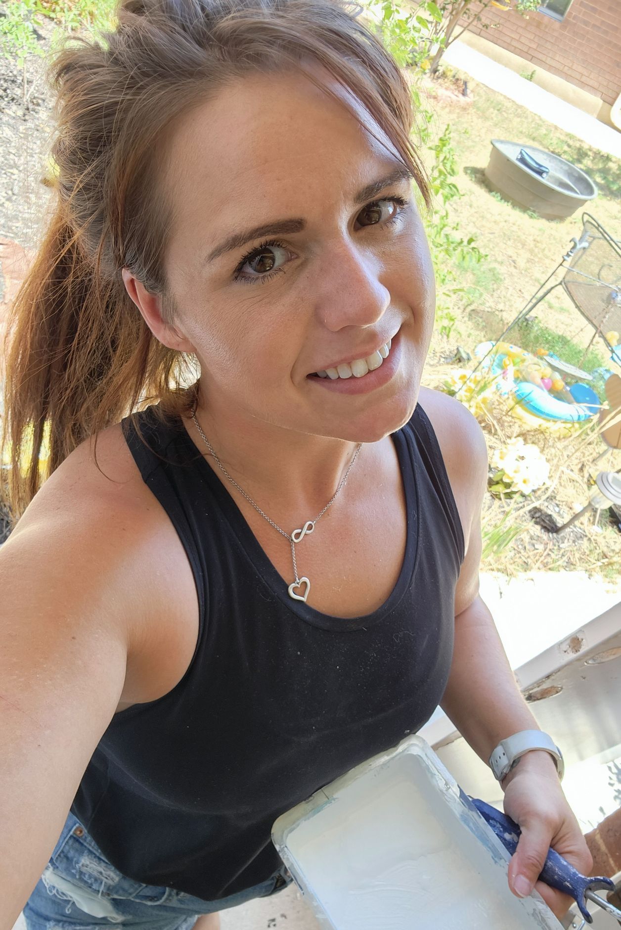 Woman with brown hair in a ponytail smiles, wearing a black tank top, holding a paint roller outdoors.