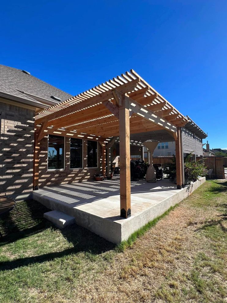 A wooden pergola attached to a brick house, shading a concrete patio. Bright blue sky overhead.