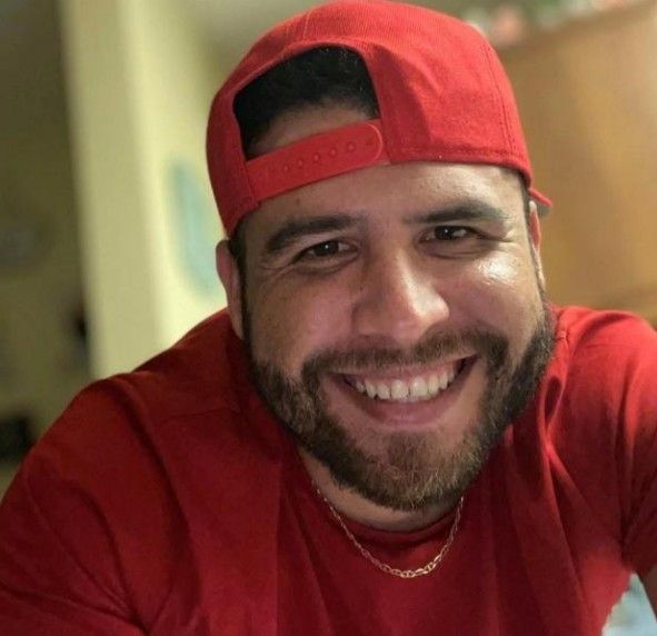 Family posing outdoors: father with a child on his shoulders, pregnant mother, and another child; near a stream.