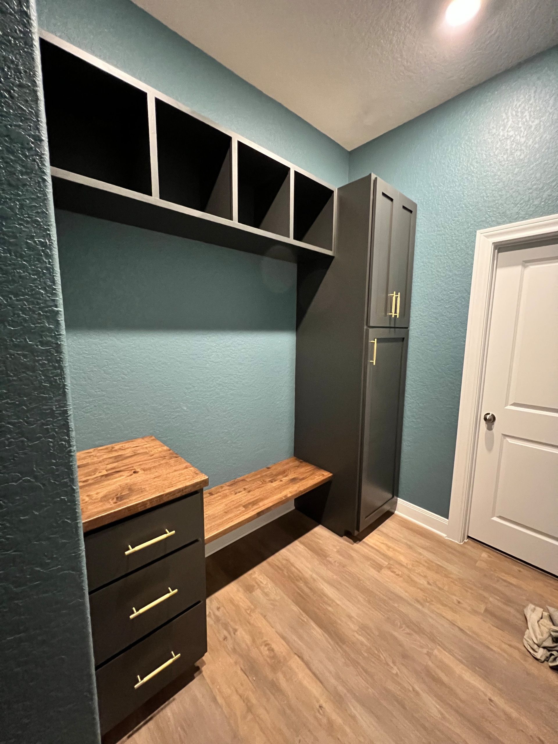 A mudroom with teal walls, dark gray built-in storage, a wooden bench, and a wood-look floor.