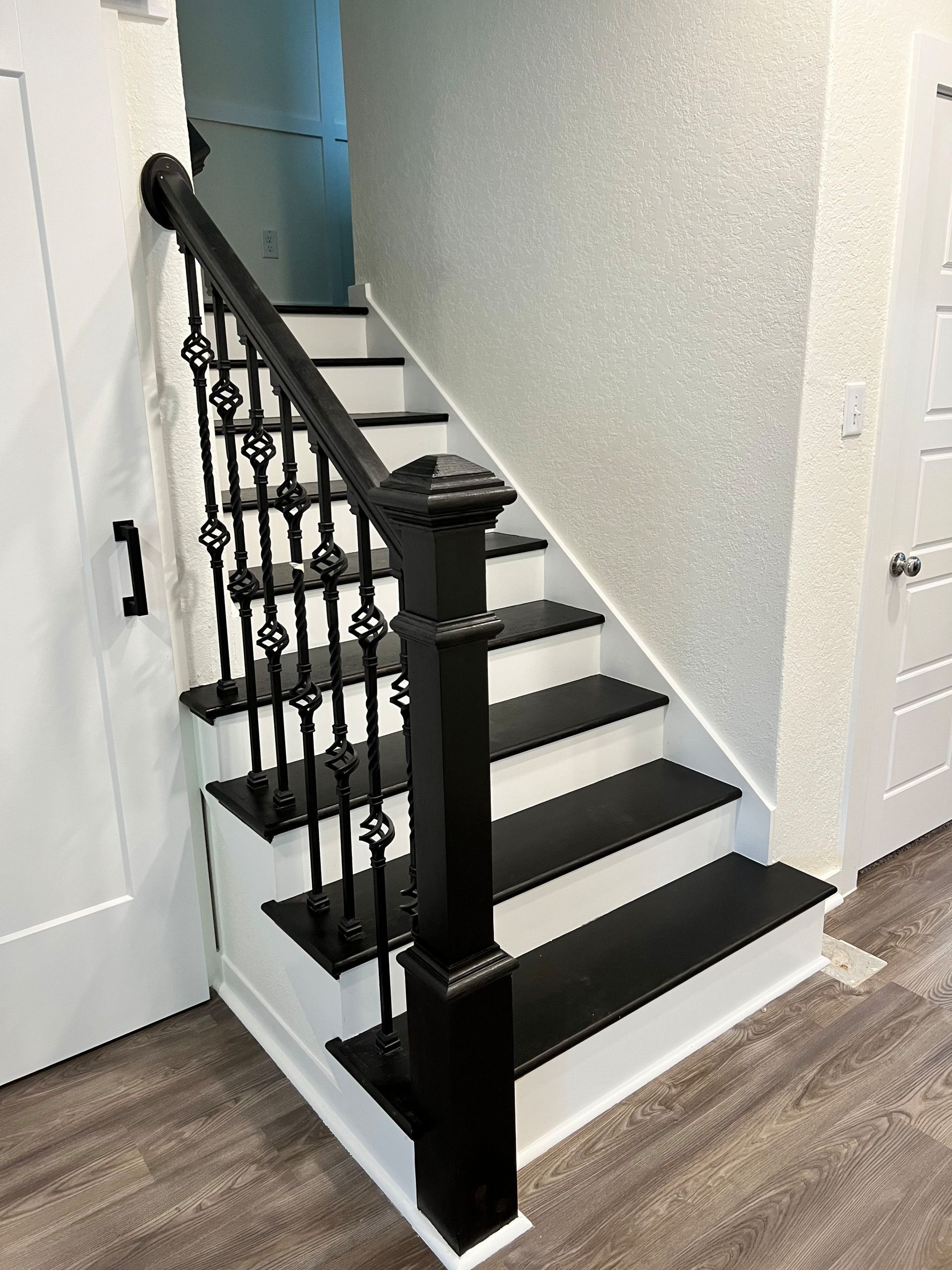Staircase with black banister and railing, black treads, and white risers. Located near a white door and textured white wall.