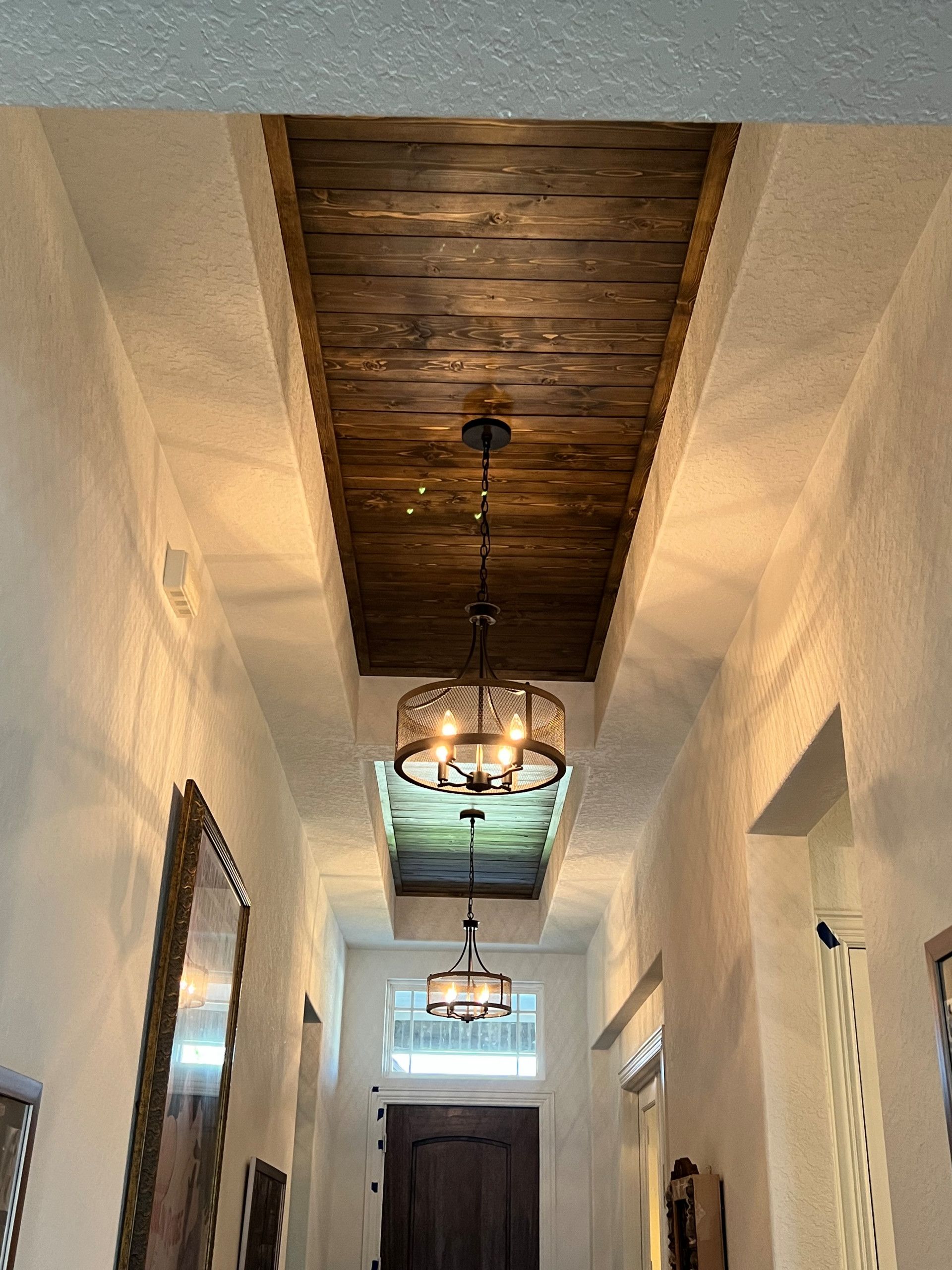 Hallway with a dark wood ceiling, white walls, and three ornate chandeliers. A dark wooden door is at the end.