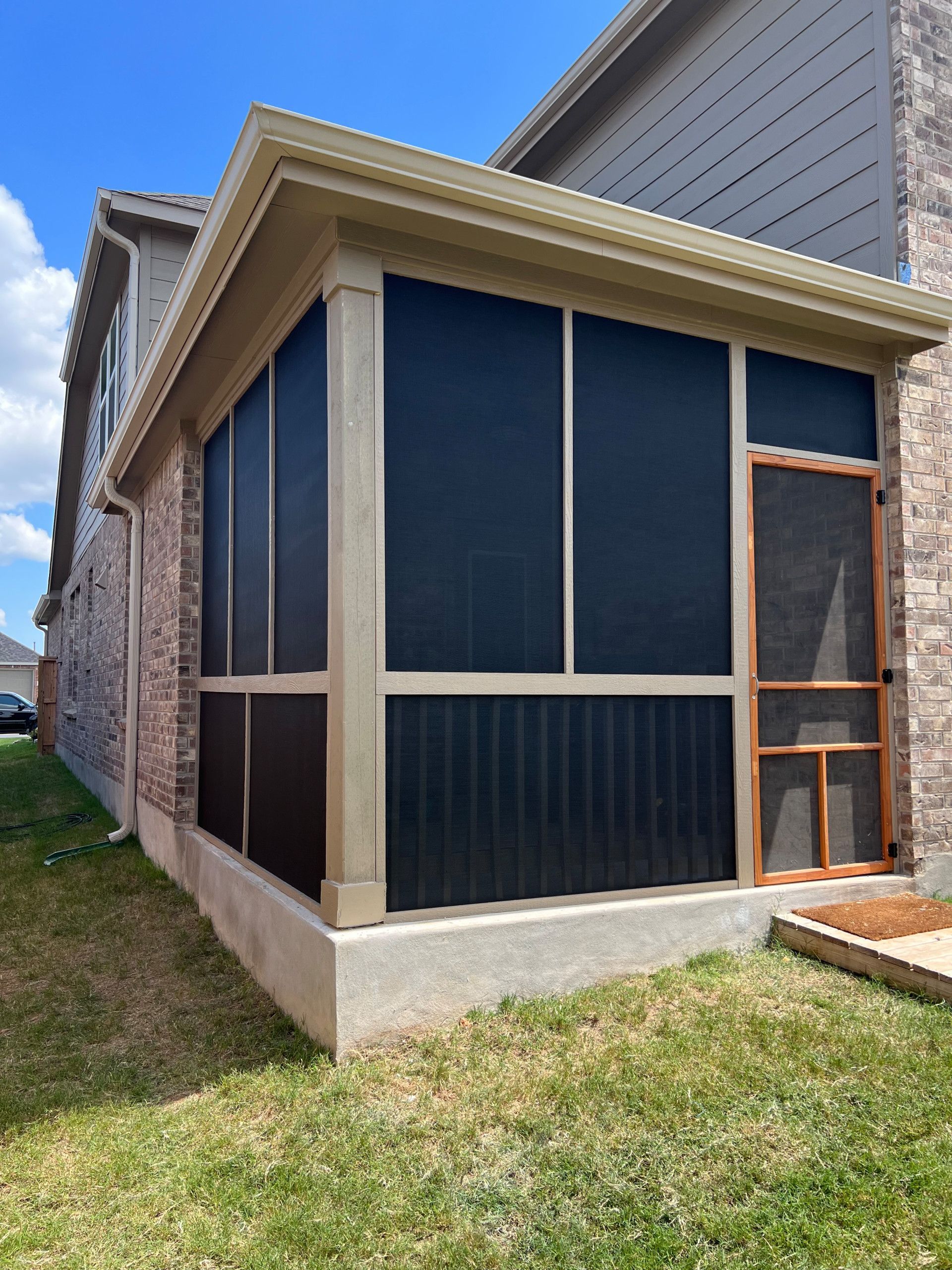 A screened-in porch attached to a two-story brick house. It has dark screens, beige trim, and a concrete base.