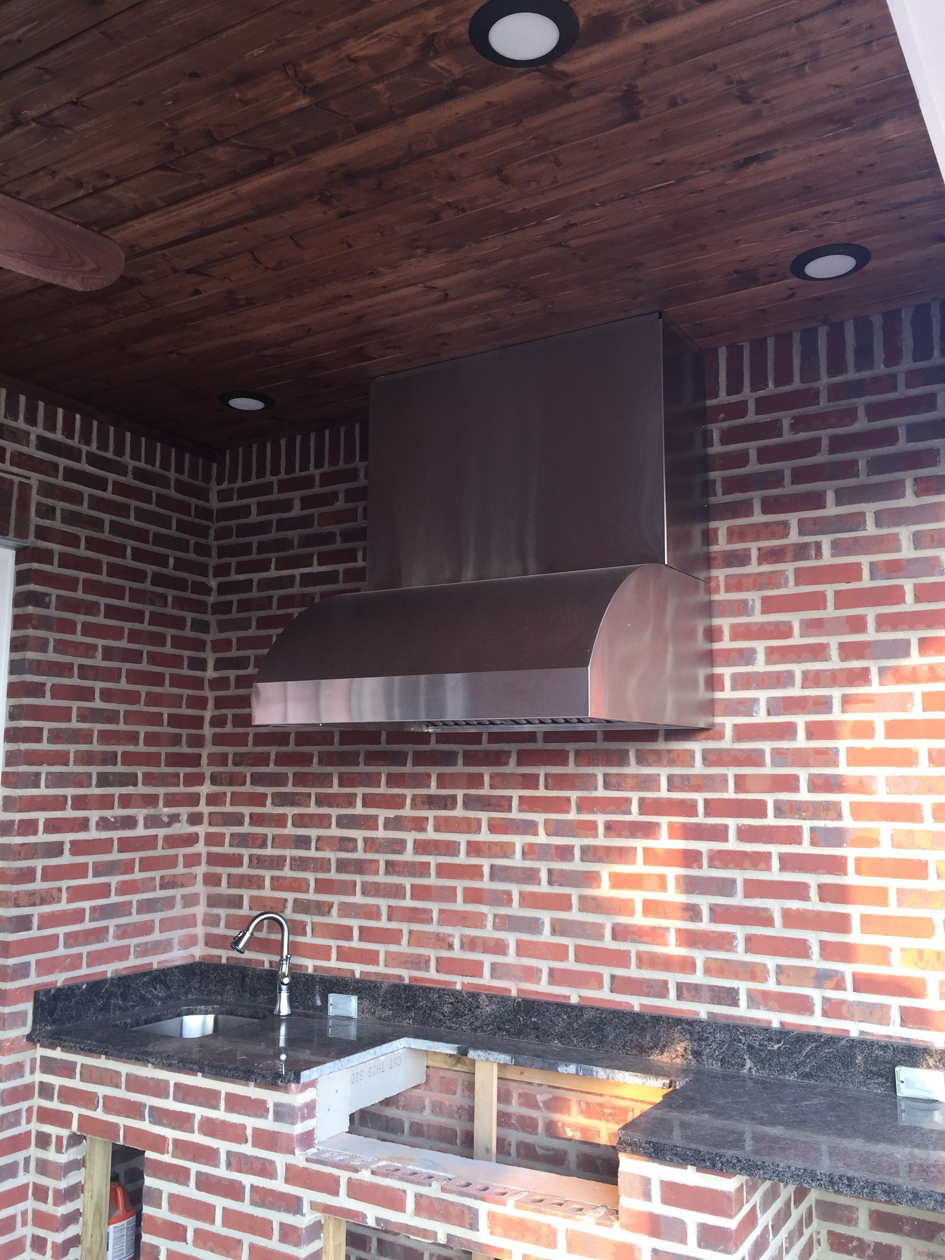 Outdoor kitchen with stainless steel range hood against a red brick wall, dark granite countertop, and wood ceiling.