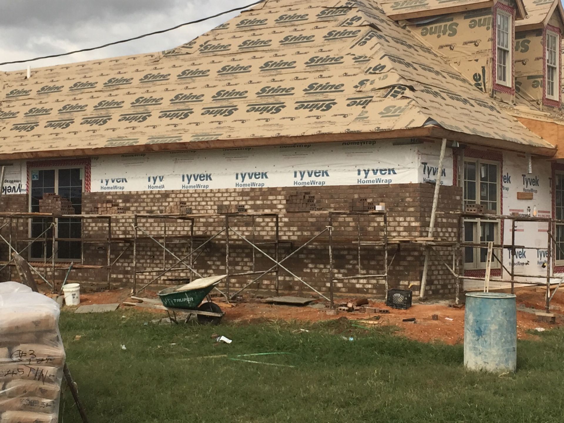 Construction site: brick siding being added to house. Scaffolding, Tyvek wrap, and green wheelbarrow visible.