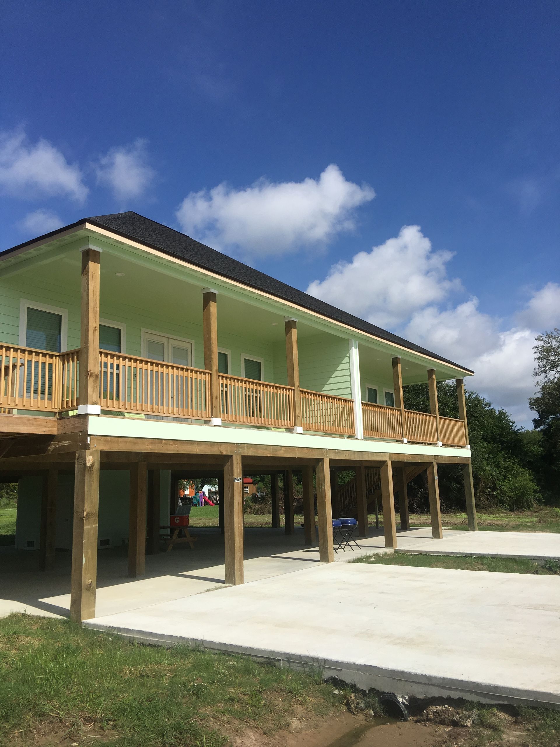 Two-story, pale green house on stilts with a wooden deck and railing, set against a blue sky with clouds.