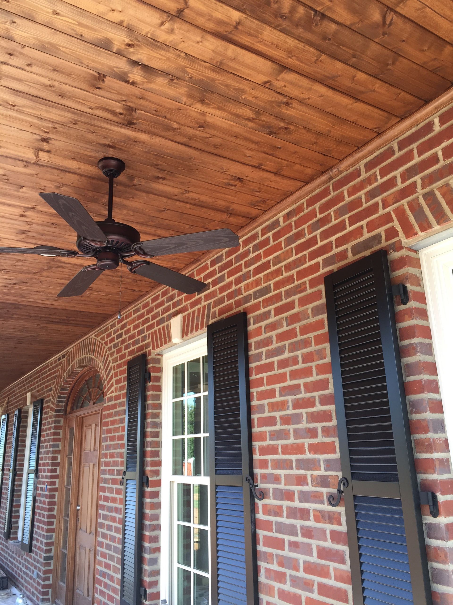 Brick porch with wooden ceiling and a dark ceiling fan; black shutters on windows.