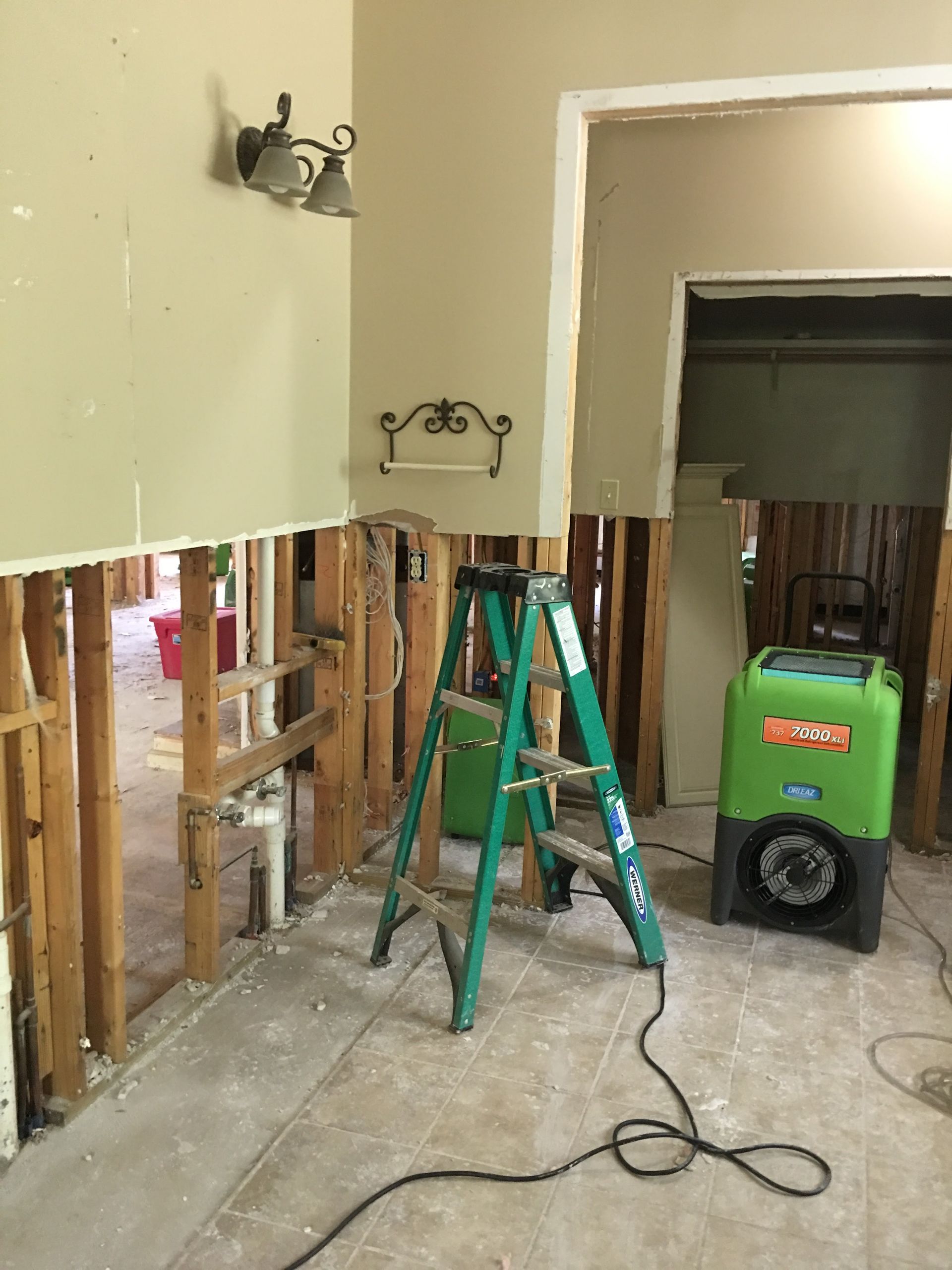 Interior room damaged by flood with exposed studs, green ladder, and dehumidifier.