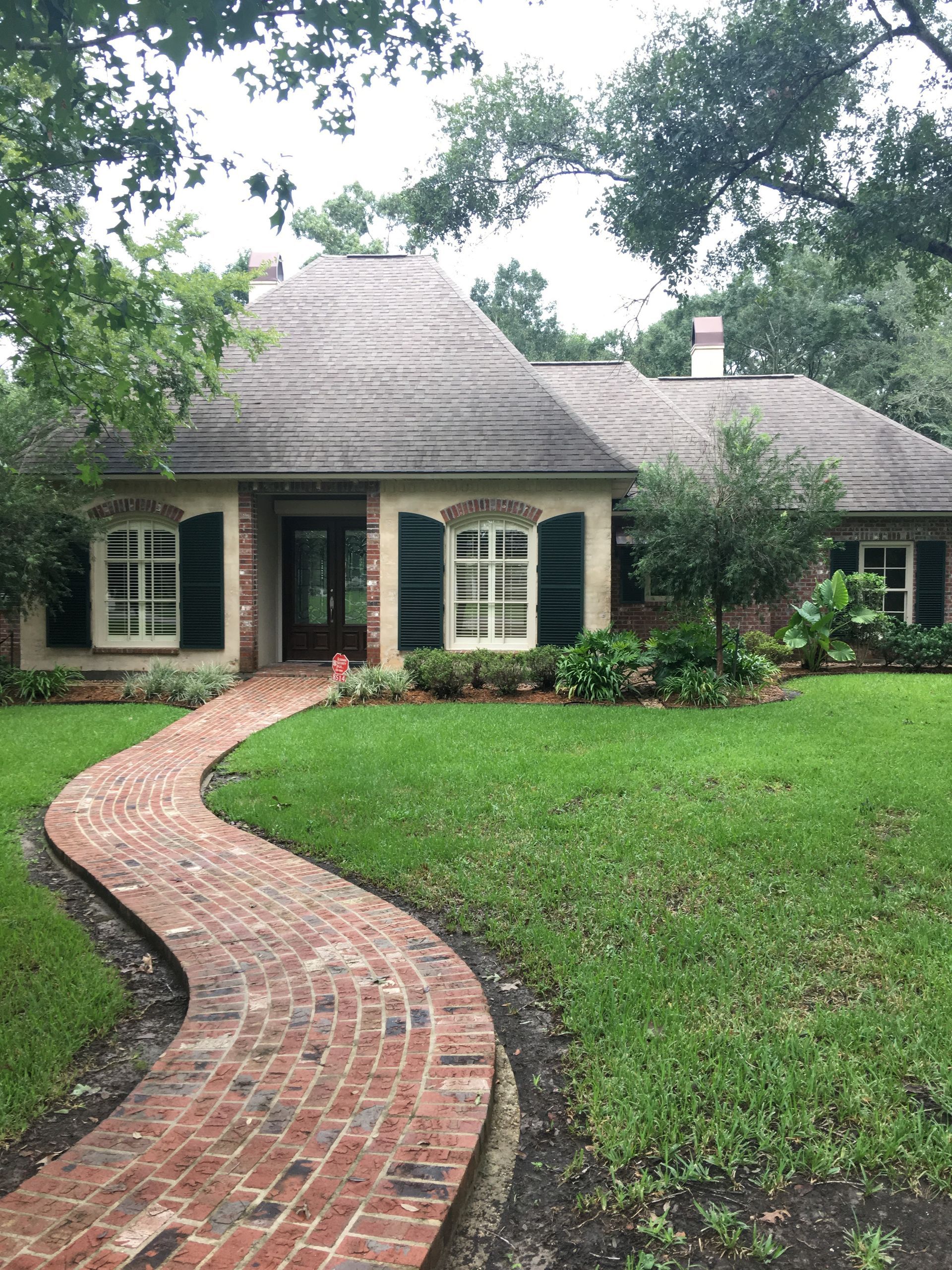 Brick pathway leads to a charming house with a thatched roof, green shutters, and lush landscaping.