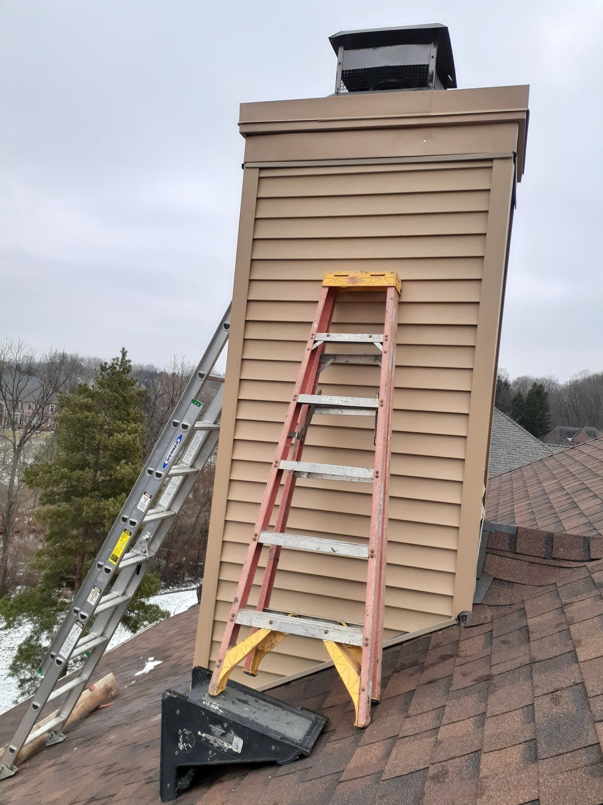 Two ladders on a brown roof lean against a tan chimney, overcast sky.