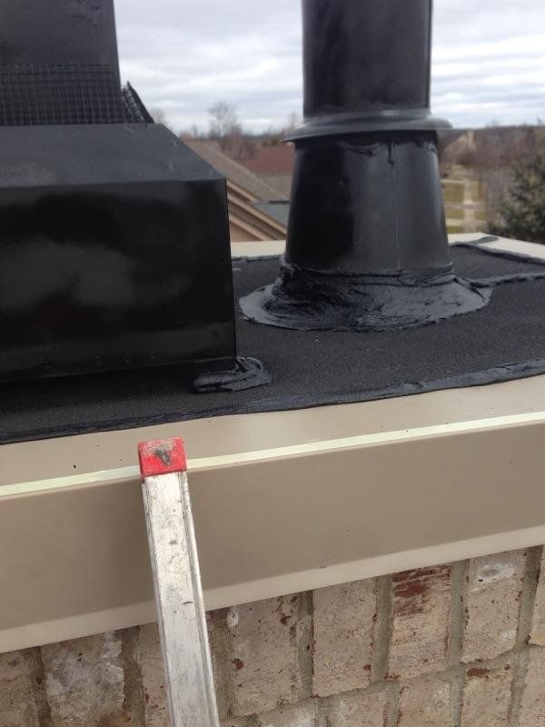 Black chimney components on a roof with a ladder leaning against a brick structure.