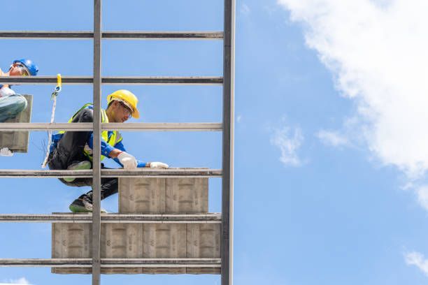 Roofing workers on metal scaffolding under a blue sky, wearing safety gear and handling materials