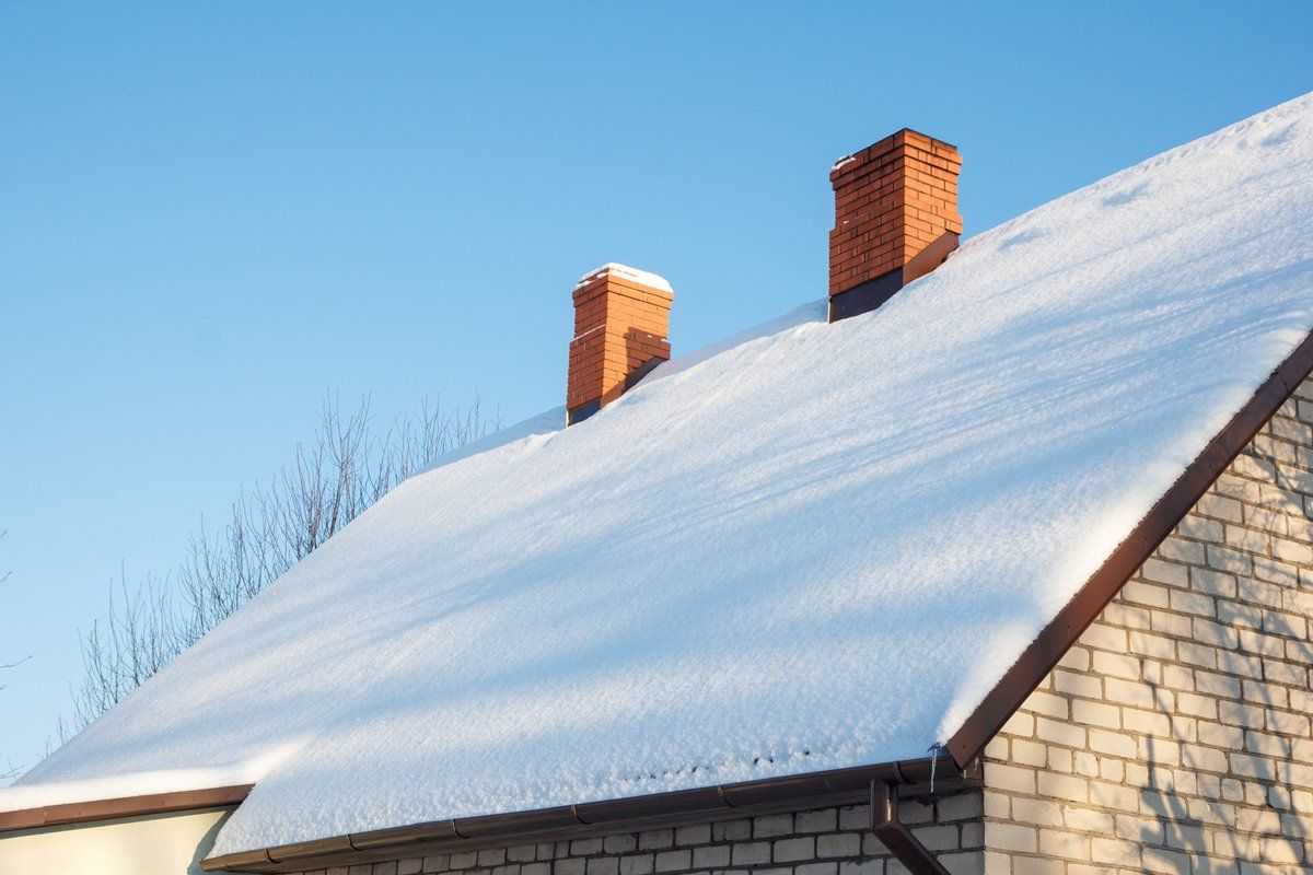 Snow-covered house roof with two brick chimneys against a clear blue sky.