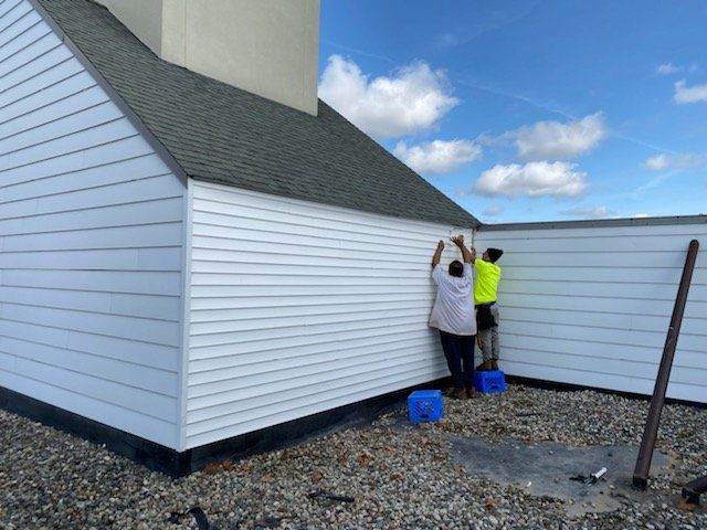 Two people installing white siding on a building's roof. Blue sky, gray roof and gravel.