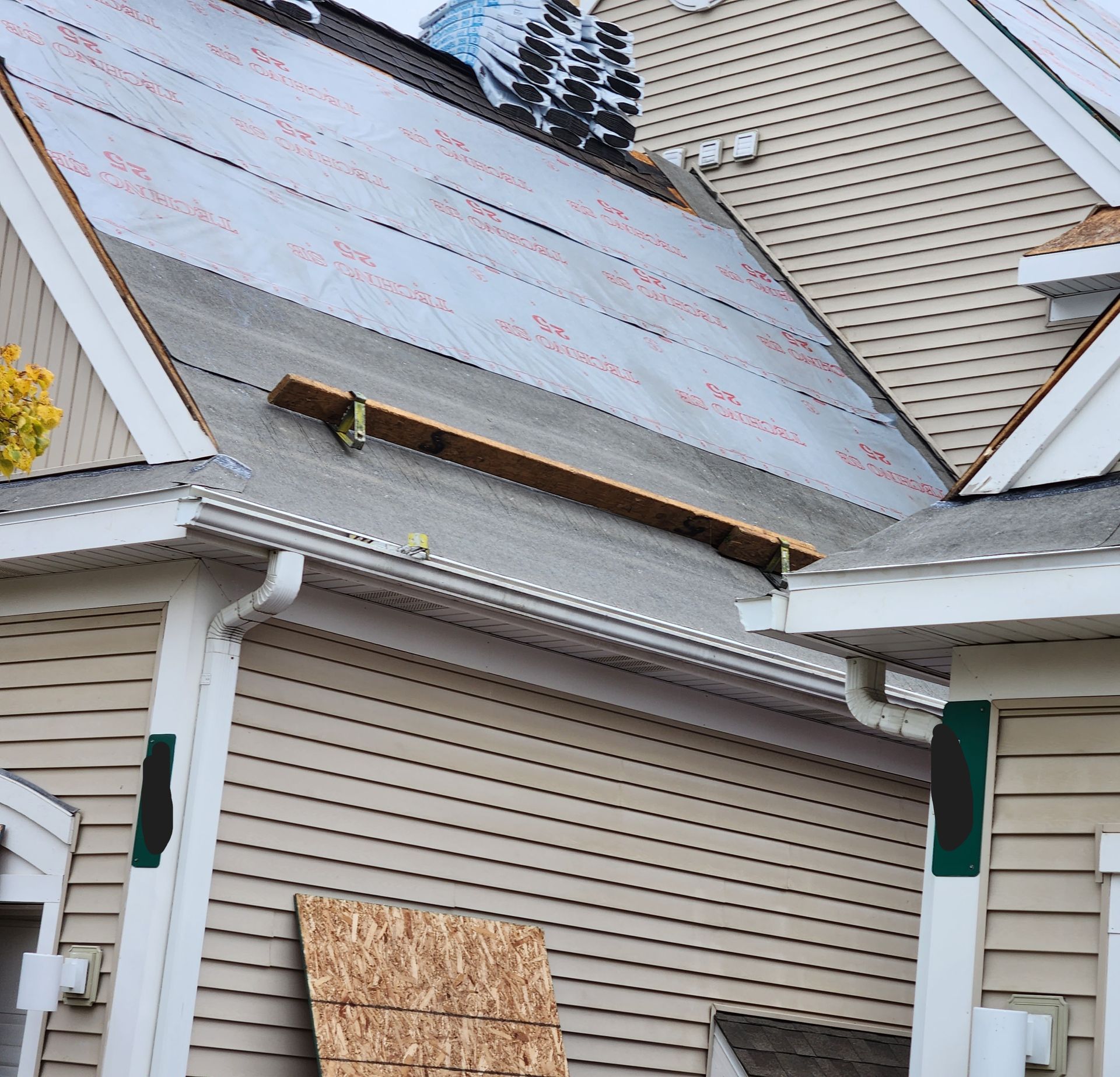 Roof under construction with exposed underlayment, shingles, and wooden plank, beside beige siding.