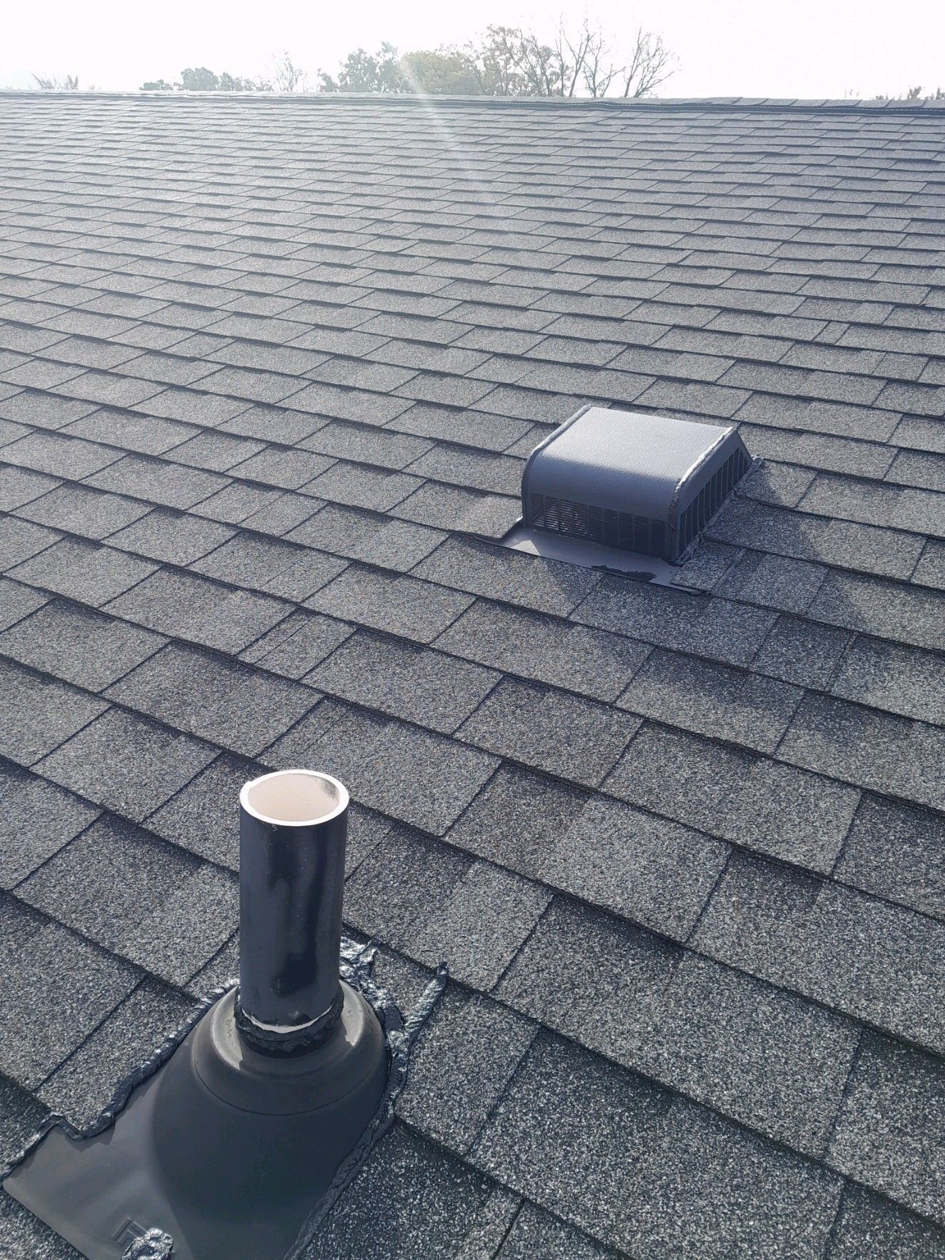 Black asphalt shingle roof with two vents: a rectangular one and a round pipe.