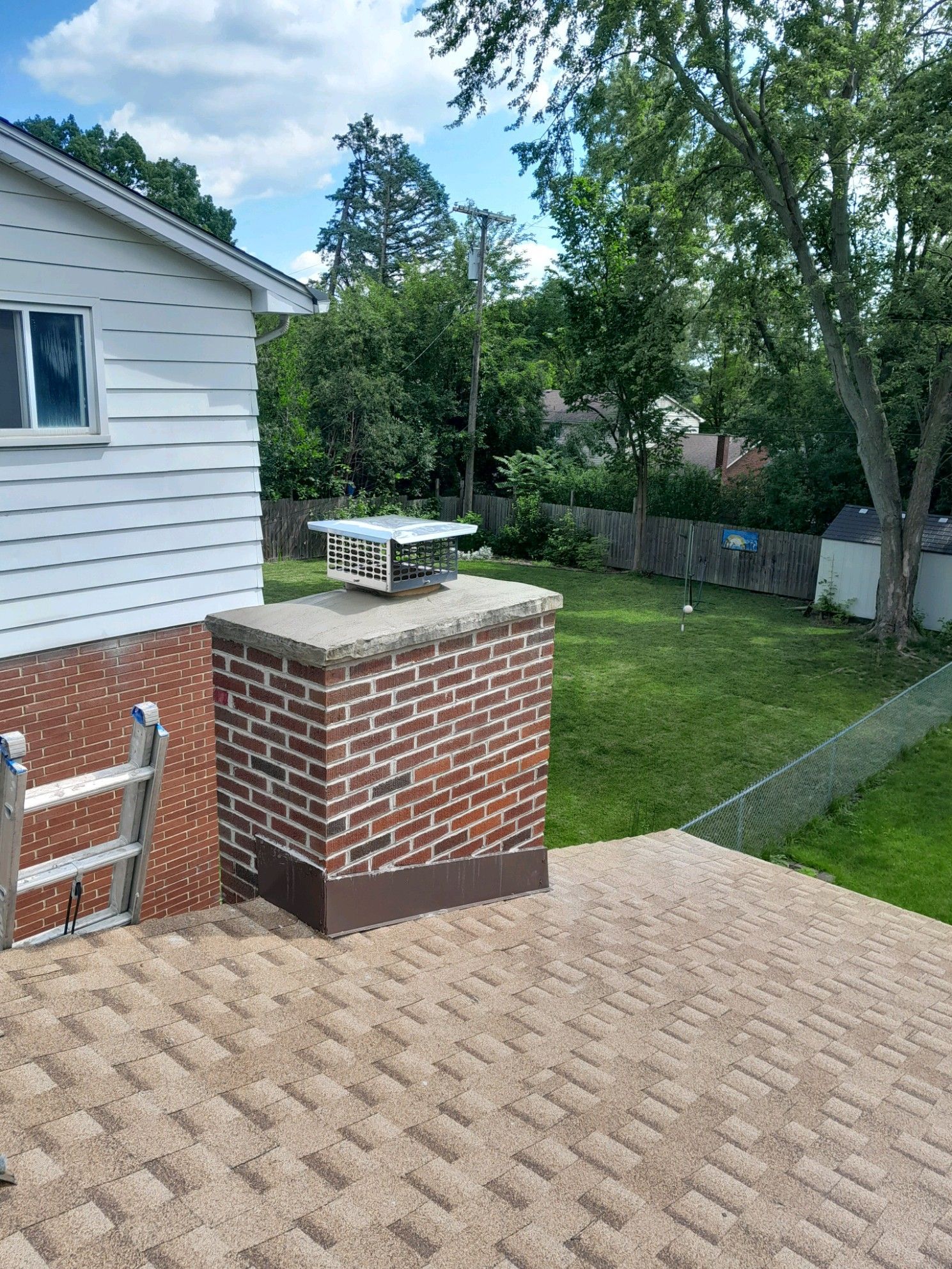 Brick chimney on a shingled roof, with a white house and green yard in the background.
