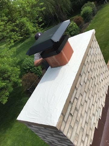 Chimney on a roof with a person installing a black chimney cap, green trees in the background.