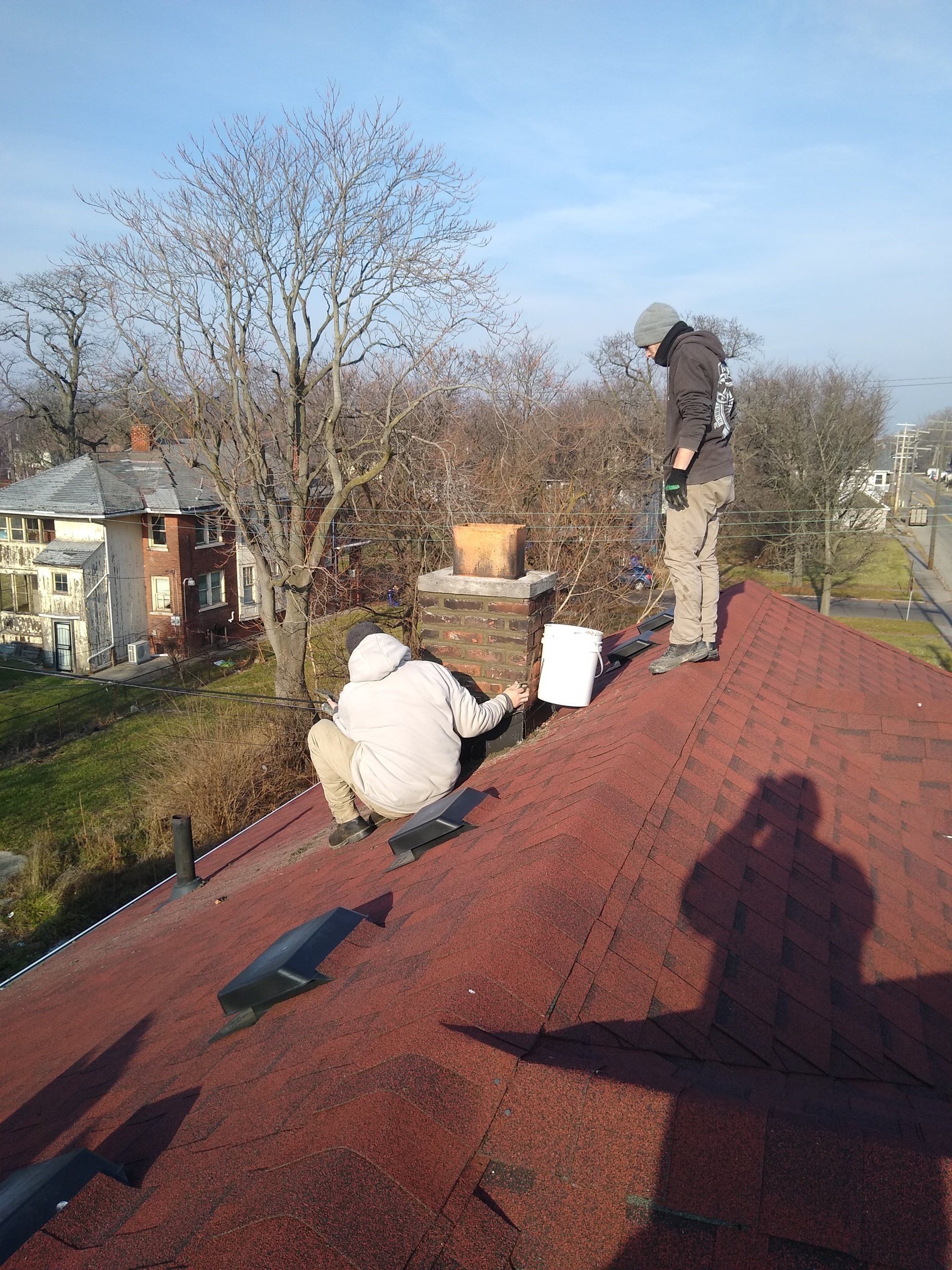 Two workers on a red shingled roof near a brick chimney, one looking at paperwork.