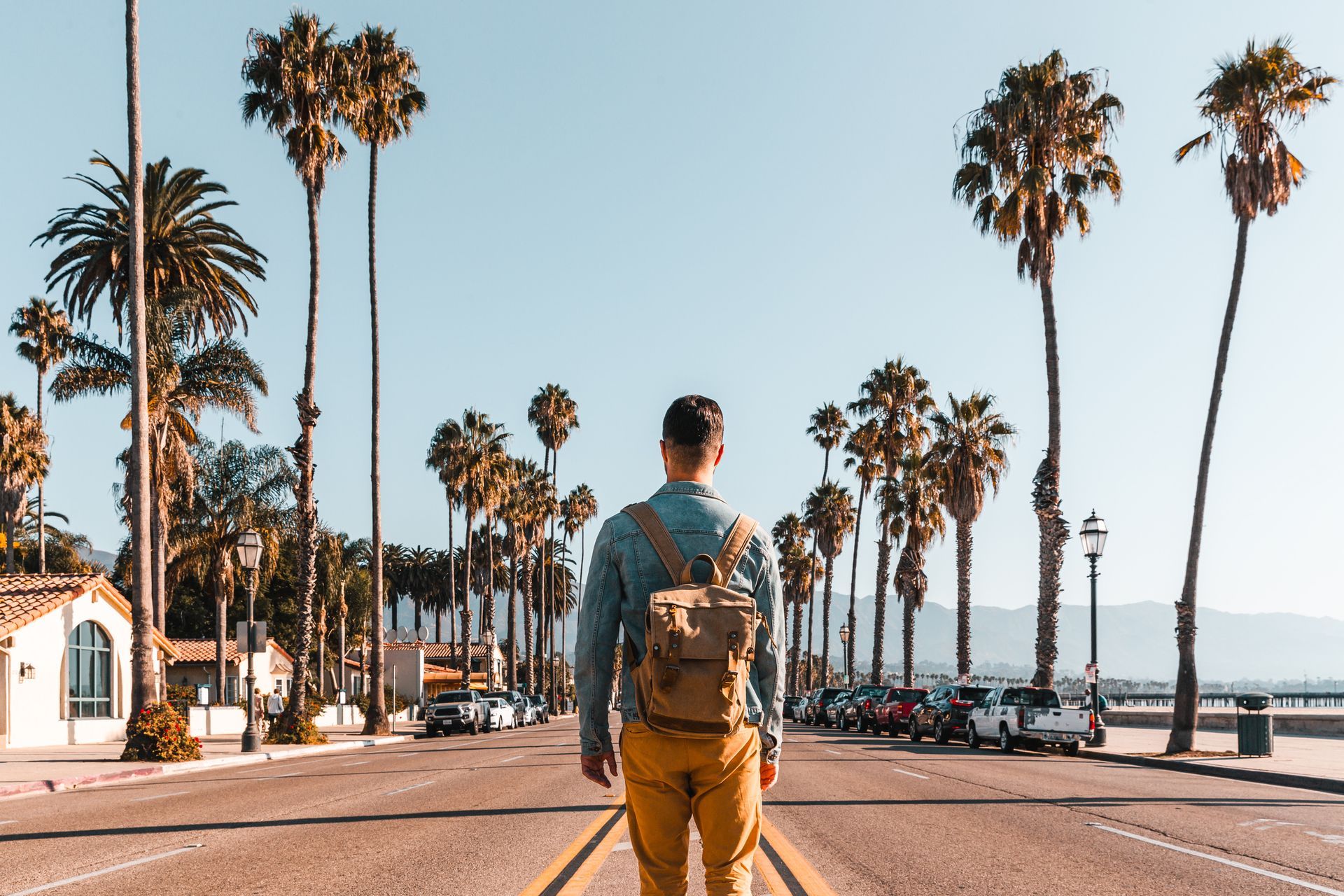 A man with a backpack is walking down a street with palm trees in the background.