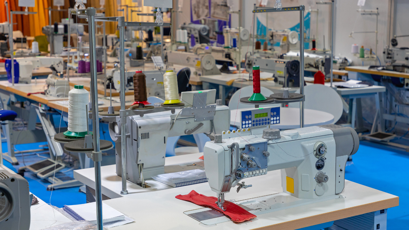 Industrial sewing machines with colorful spools of thread on white workbenches in a factory setting with a blue floor.