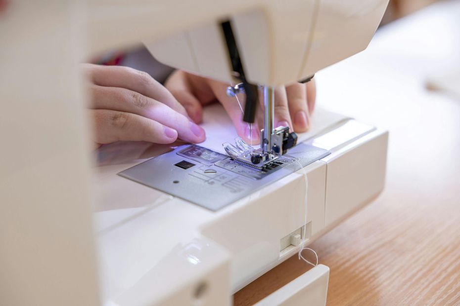 Hands guide white fabric under the needle of a white sewing machine on a wooden table.