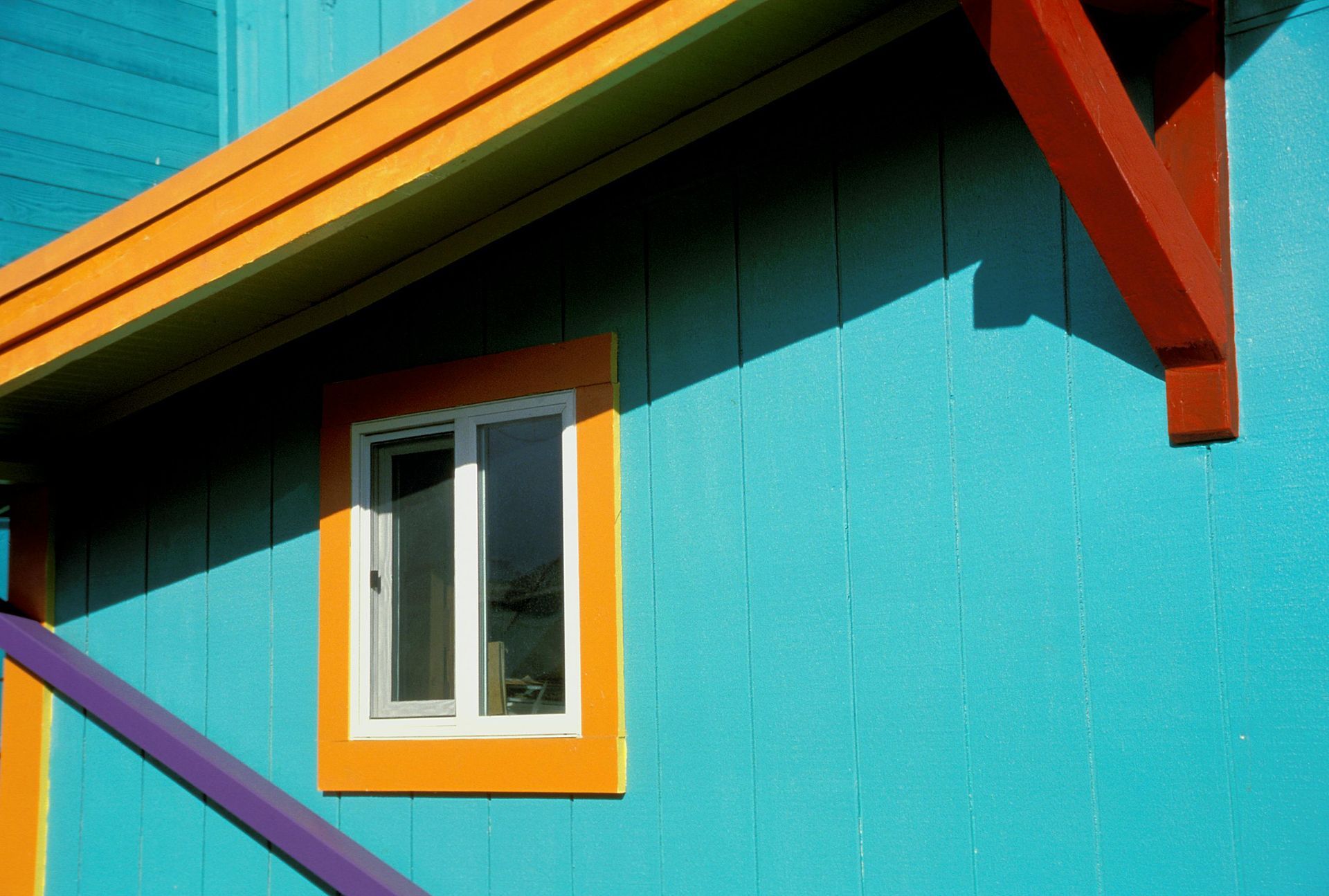 Bright turquoise building with orange and purple accents, a white-framed window, and a red support beam.