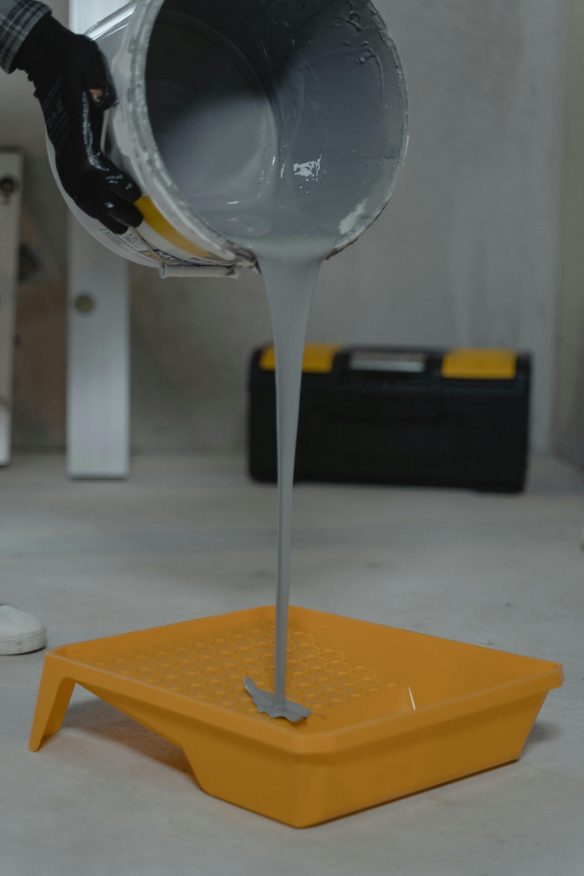 Person pouring gray paint from a bucket into a yellow paint tray, setting a DIY scene.