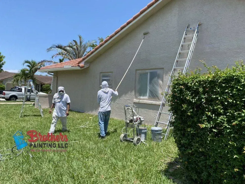 Two workers painting a house exterior, with ladders and paint equipment on the lawn.