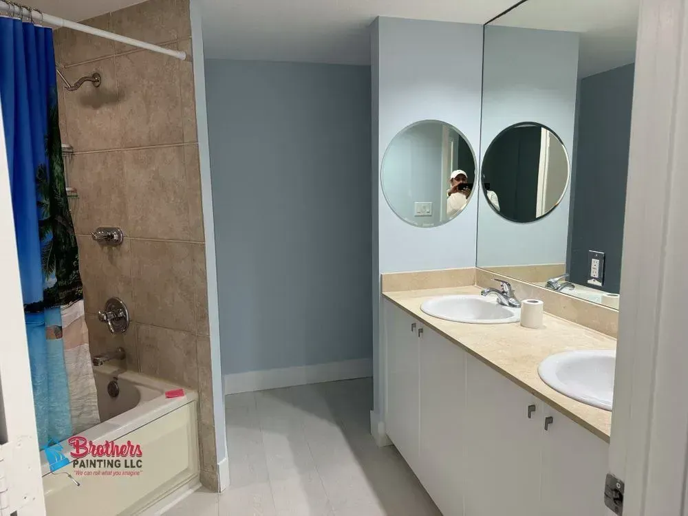 Public restroom with a shower stall, double sinks, and round mirrors in light blue and beige tones