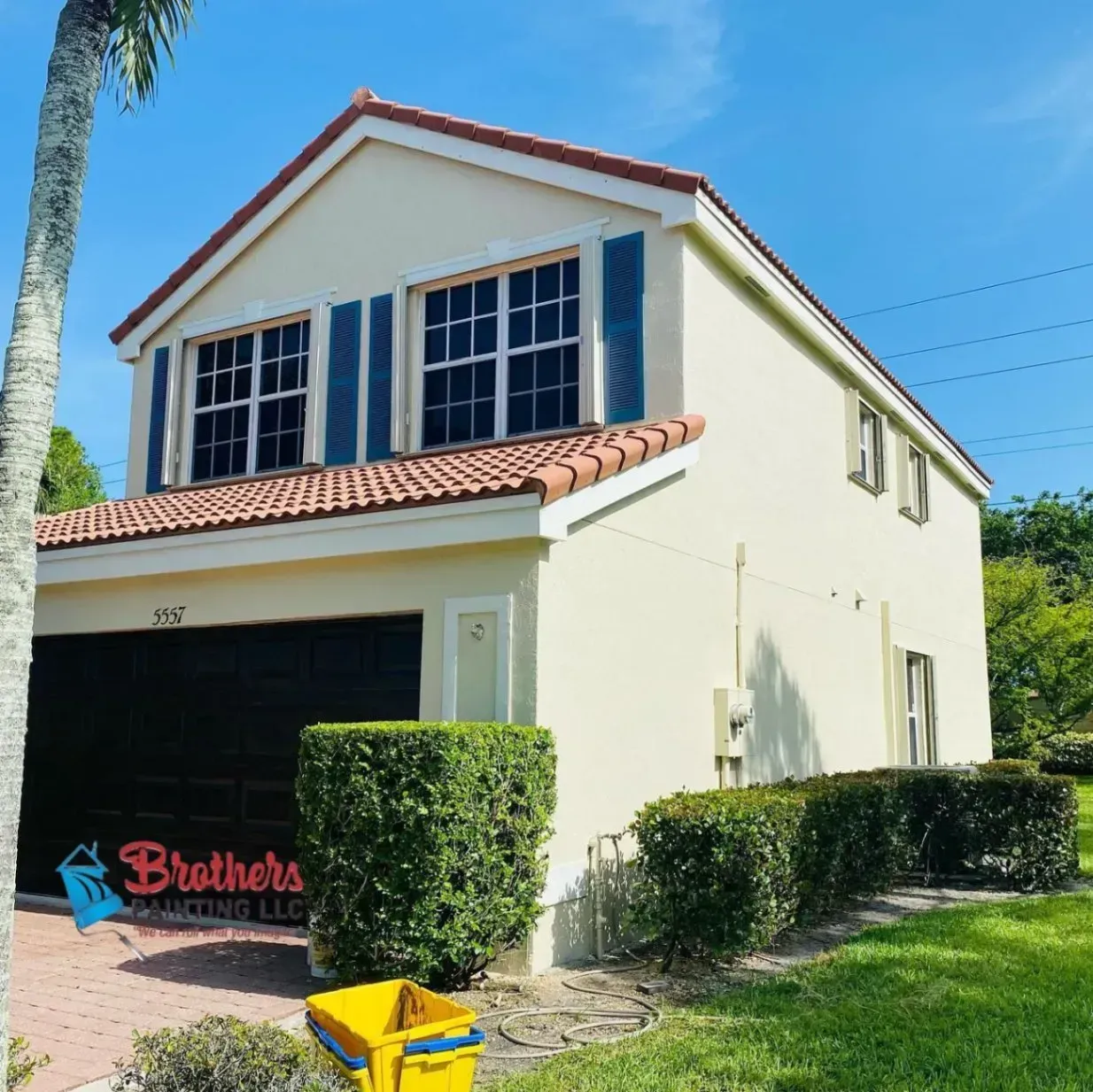 Two-story cream stucco house with red-tile roof, black shutters, and a garage in a sunny suburban yard