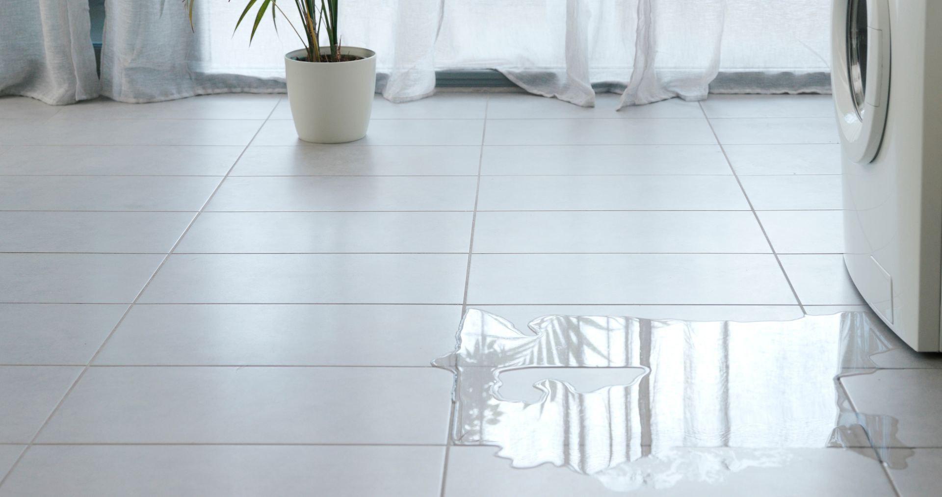 A large water leak spreading on white tiled flooring next to a washing machine.