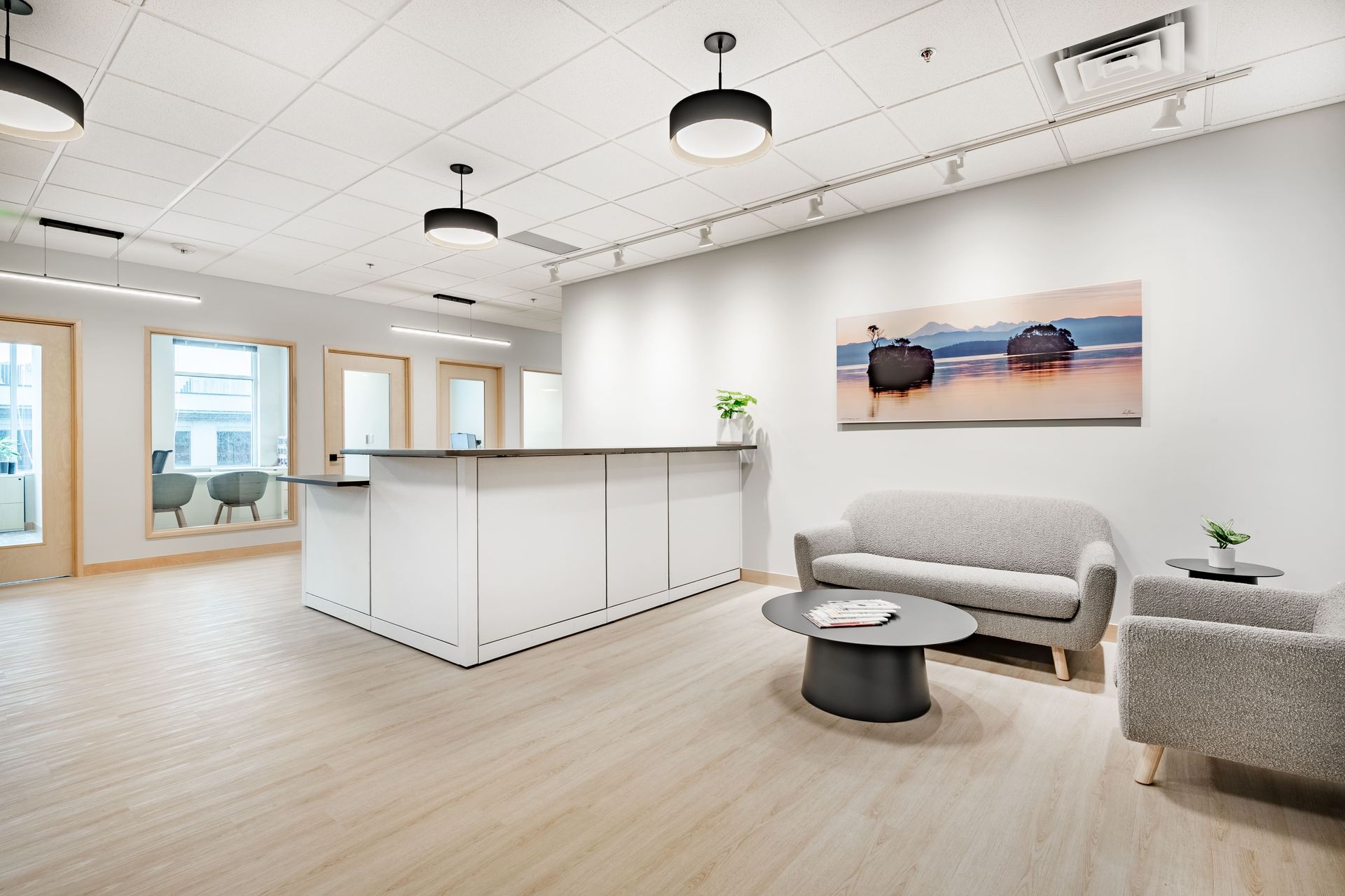 A bright office reception area featuring a white counter, gray sofa, coffee table, and a wall painting of a beach.