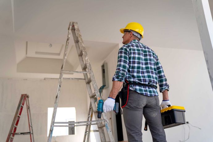 A man is standing on a ladder in a room holding a toolbox.