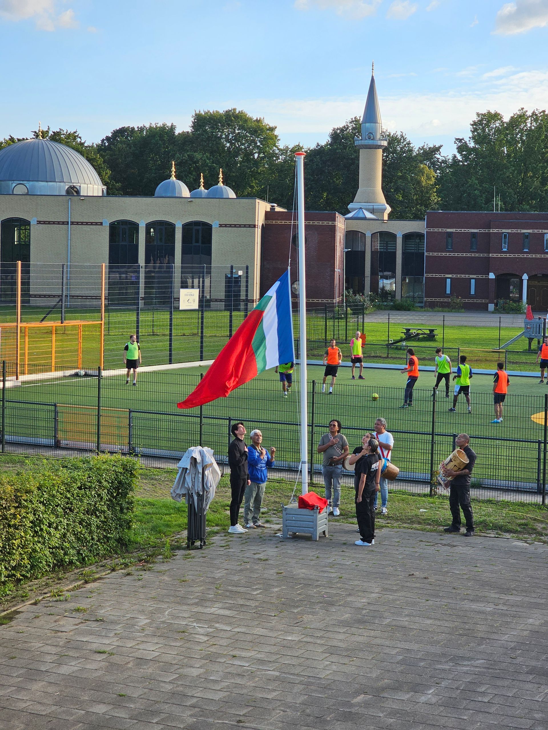 Een groep mensen staan ​​rond een vlaggenmast met een moskee op de achtergrond