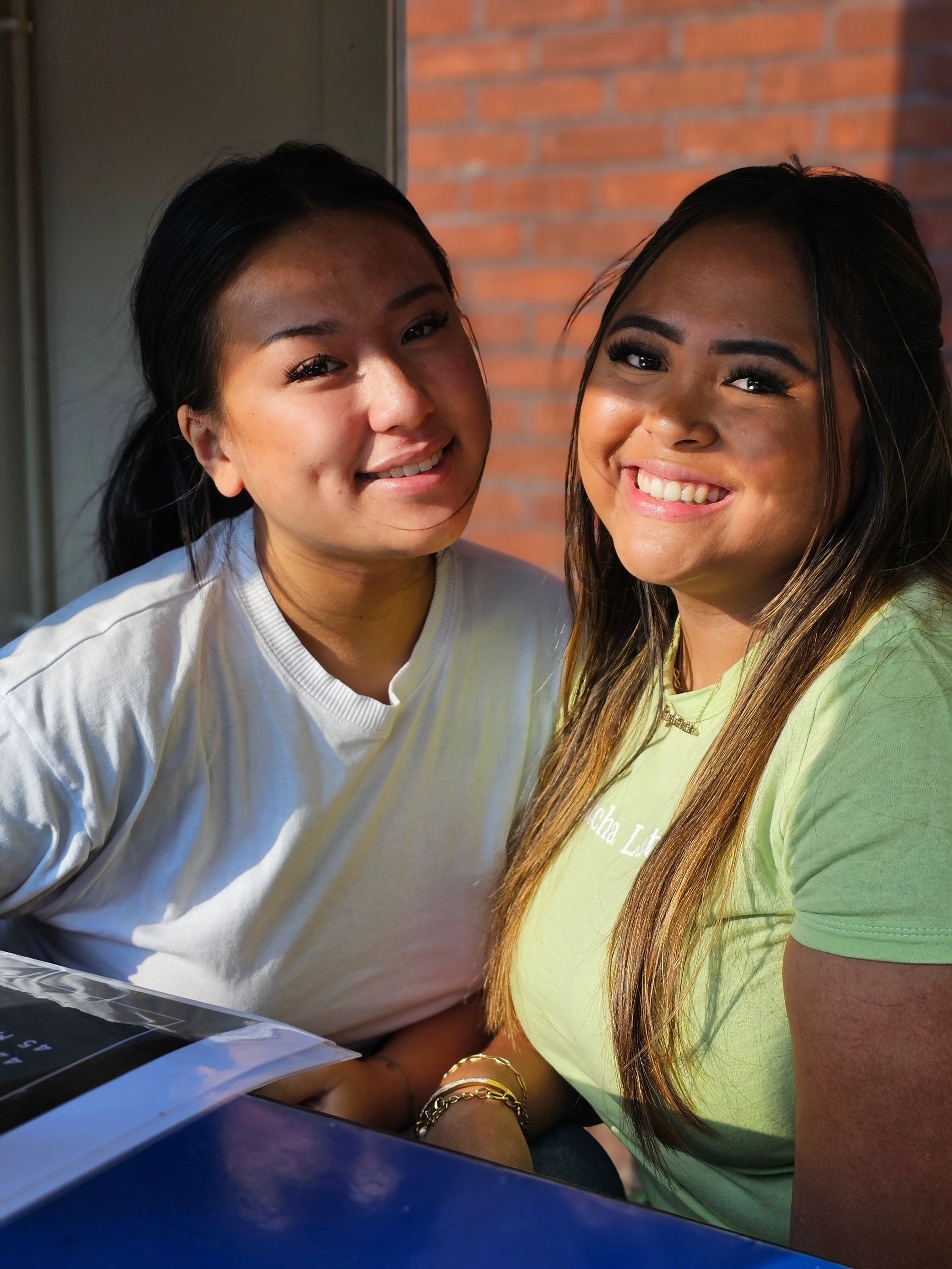 Twee vrouwen zitten naast elkaar aan een tafel en lachen naar de camera.