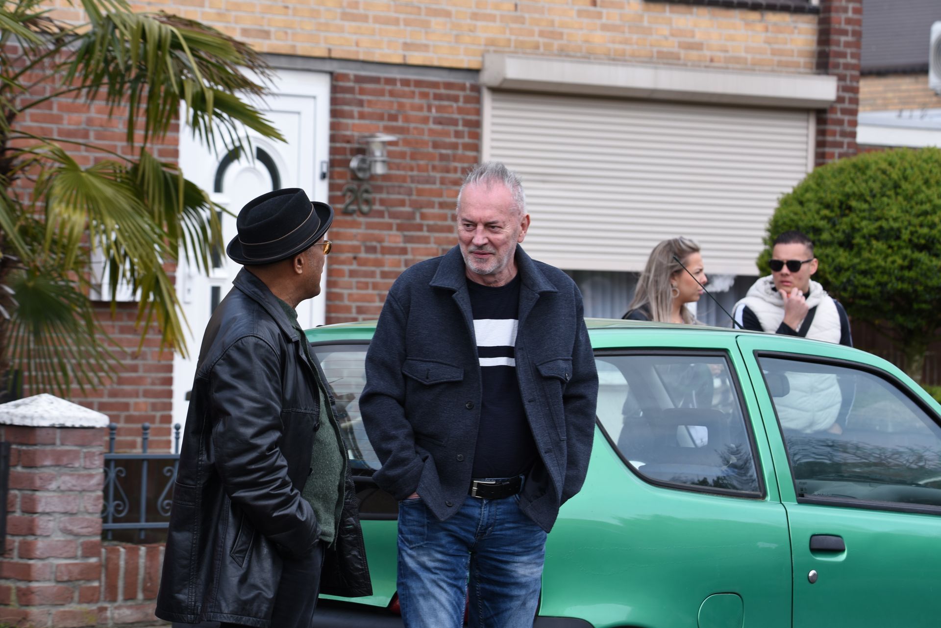 Twee mannen staan ​​naast een groene auto voor een bakstenen huis.