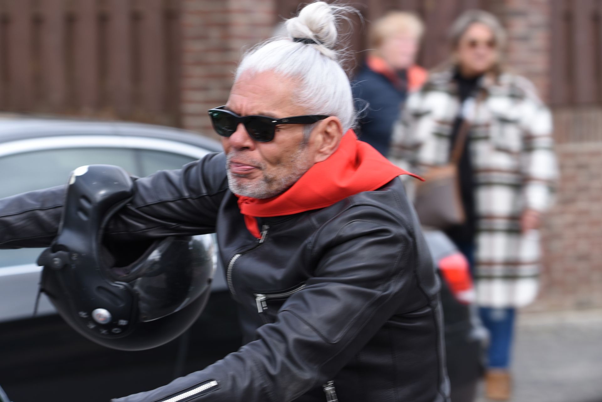 Een man met een leren jas en een rode hoodie houdt een helm vast.