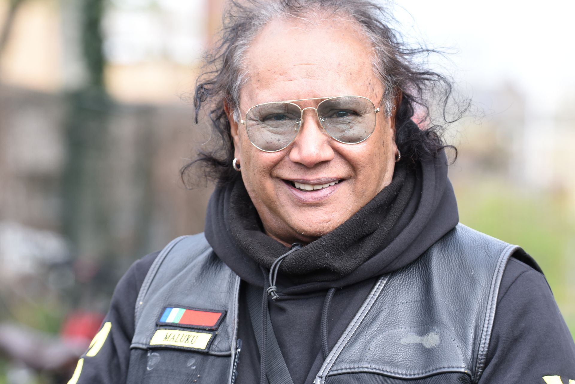 Een man met een zonnebril en een zwart vest lacht naar de camera.