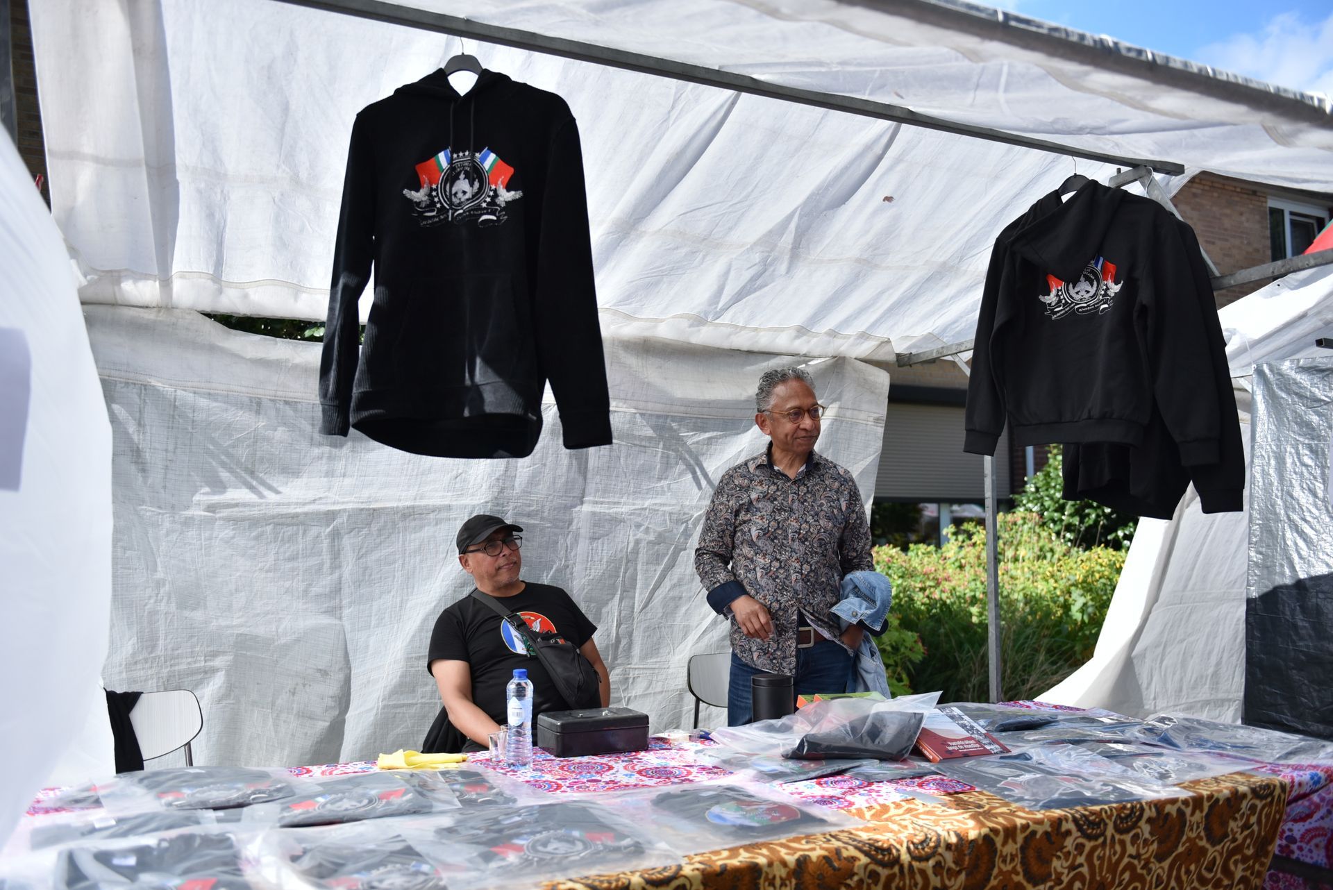Een man en een vrouw zitten aan een tafel onder een tent en verkopen overhemden.
