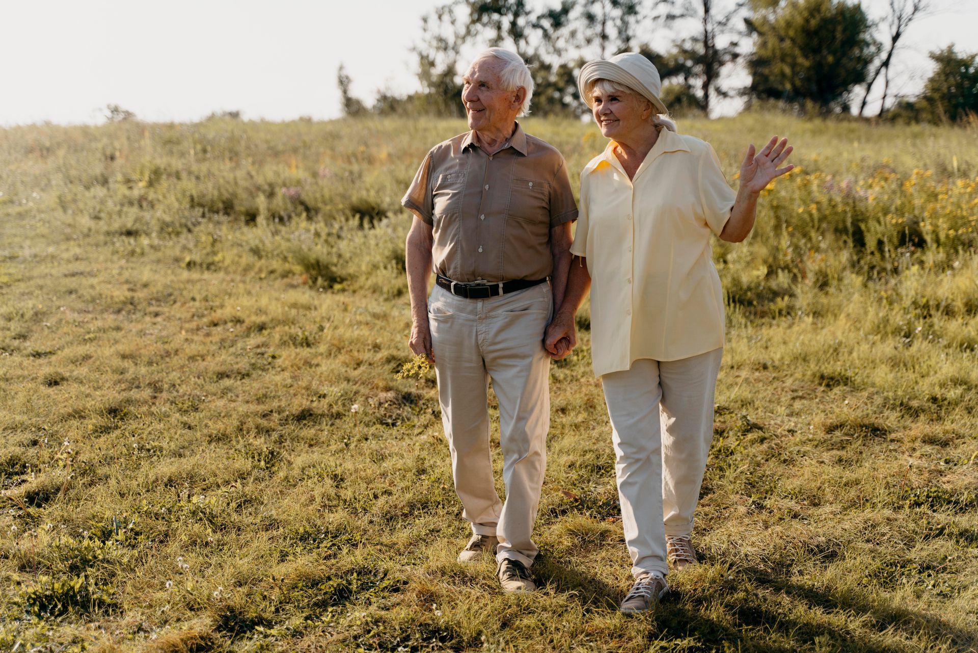 An elderly couple walks hand-in-hand across a grassy field on a sunny day. The woman waves.