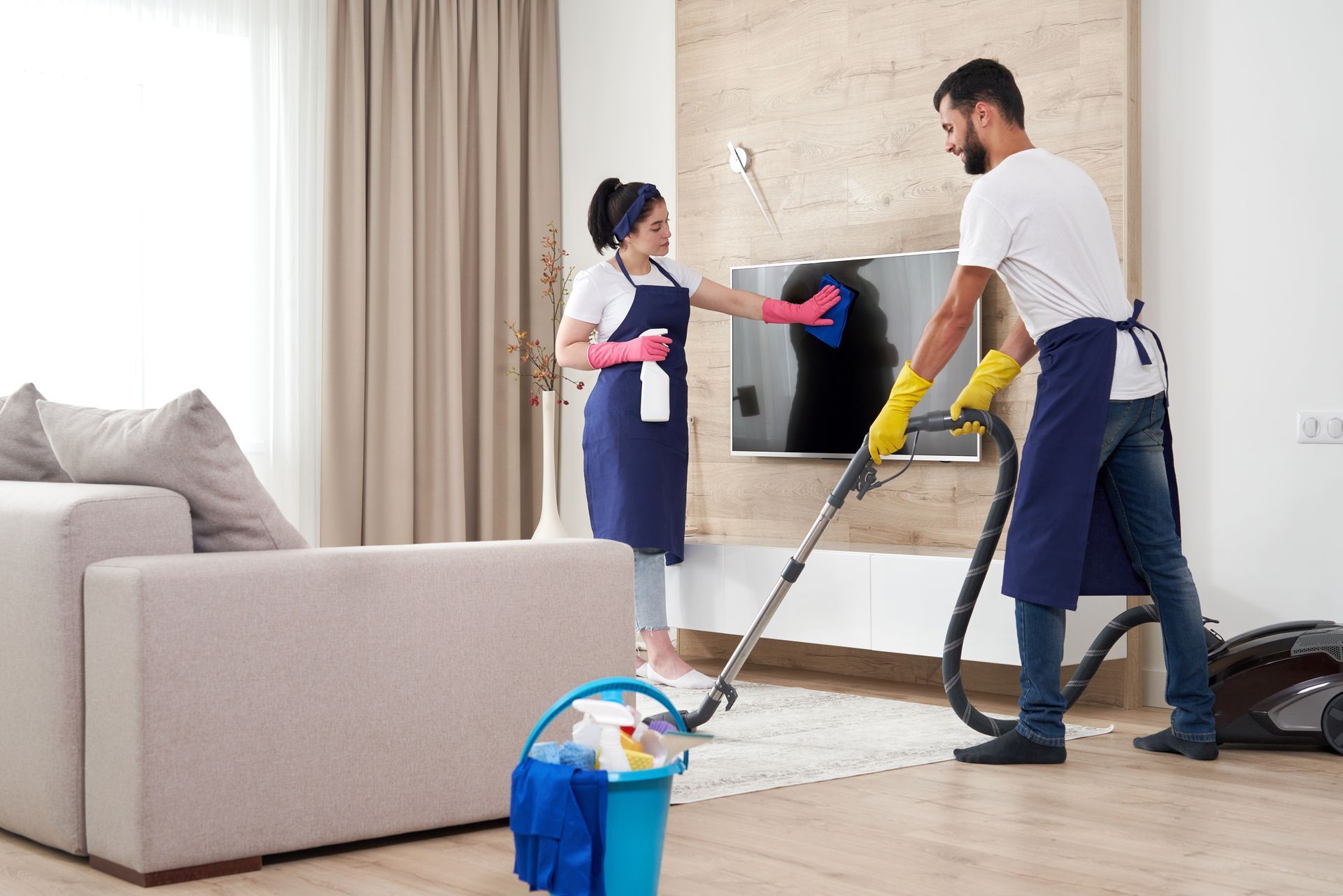 Two people cleaning a living room: one vacuums, the other dusts a TV. Both wear aprons and gloves, cleaning supplies visible.