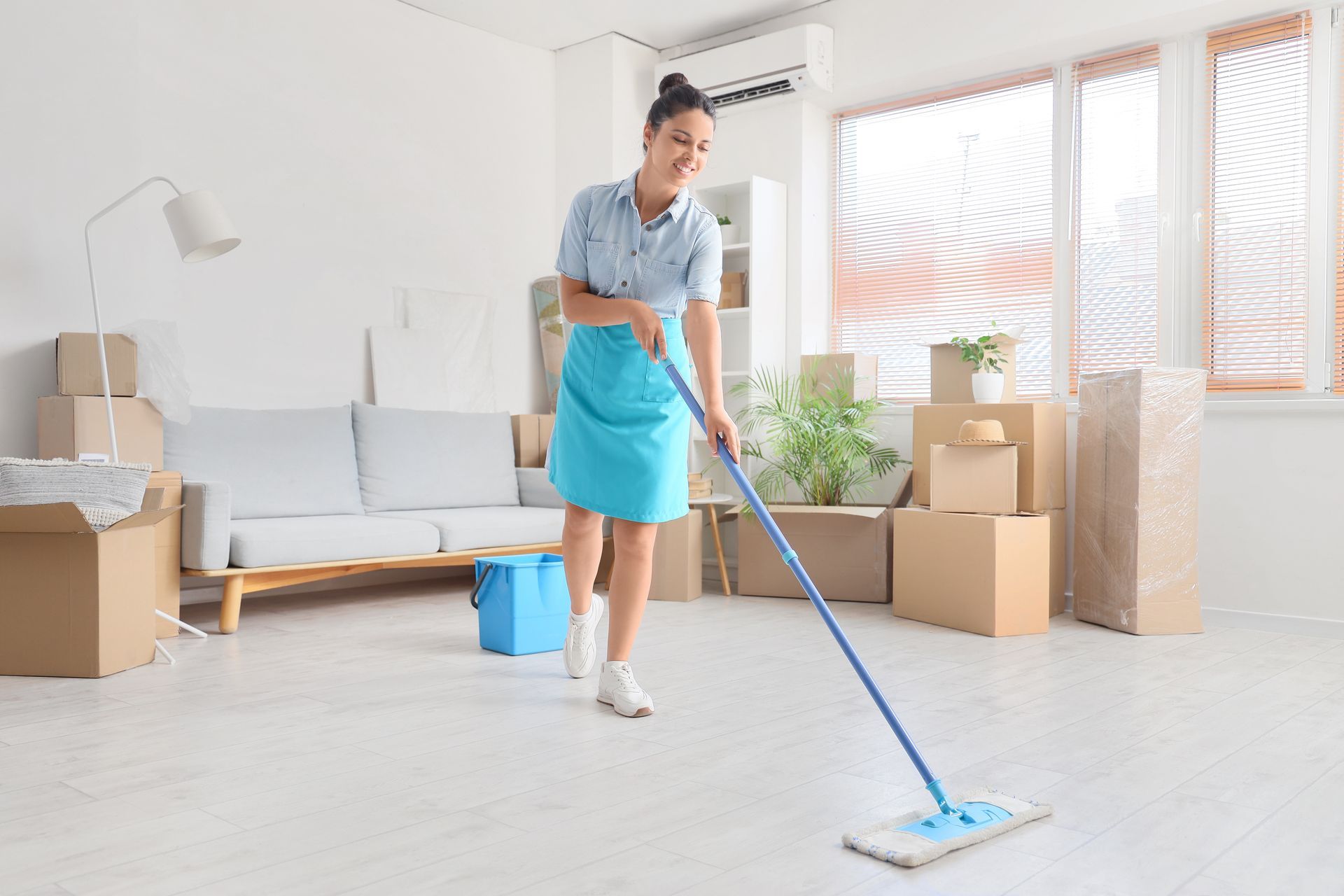 Woman mopping a room with cardboard boxes. She wears a blue skirt, smiling. Room has a sofa and windows.