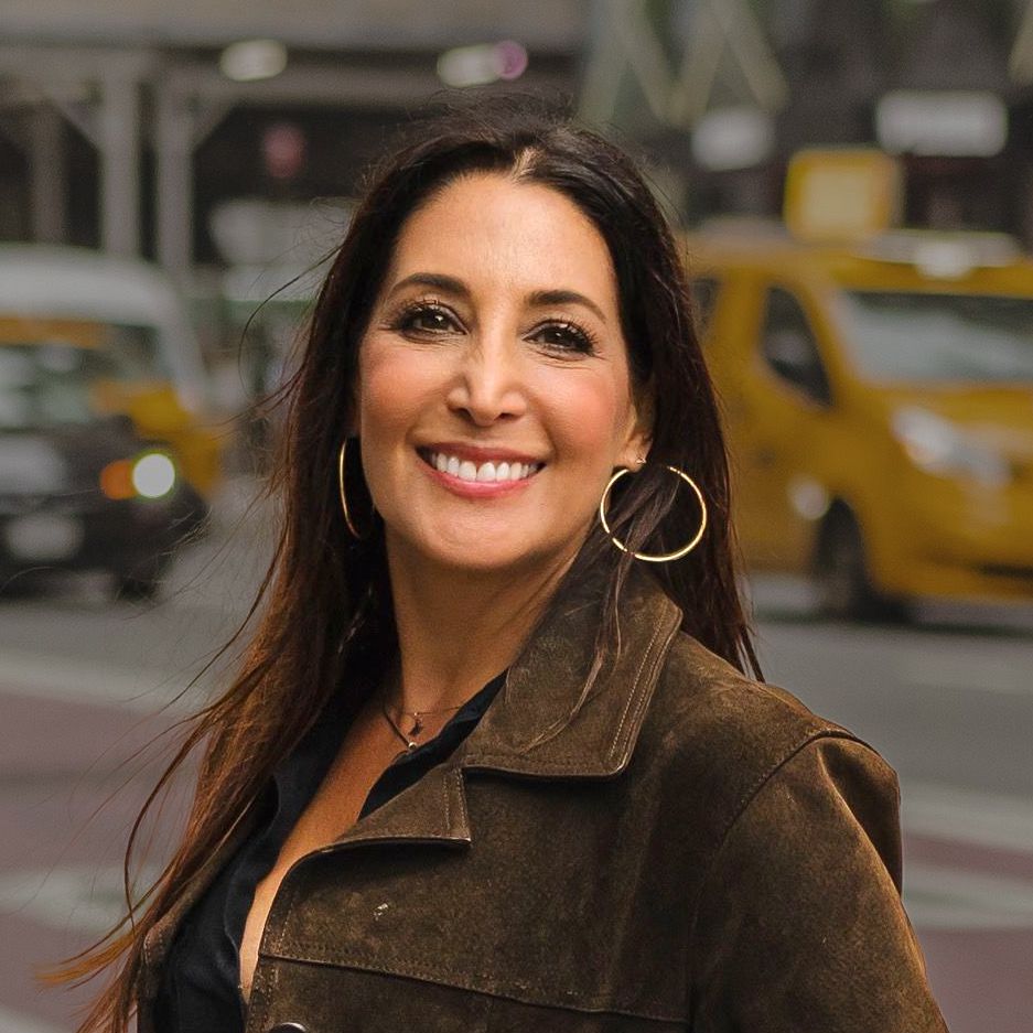 Matchmaker & Dating Coach Celine Ikeler with dark hair smiling, wearing a brown jacket, and hoop earrings, outside near taxis.