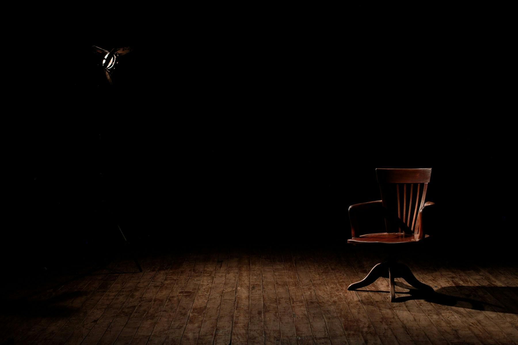Wooden chair on a stage, spotlighting an unknown object. Dark background.