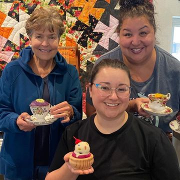 Three women are standing next to each other holding cups of tea and cupcakes.