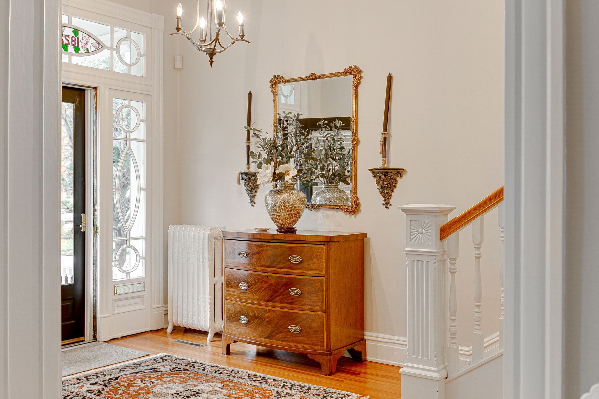 Elegant foyer with wooden dresser, ornate mirror, chandelier, stained-glass door, and staircase