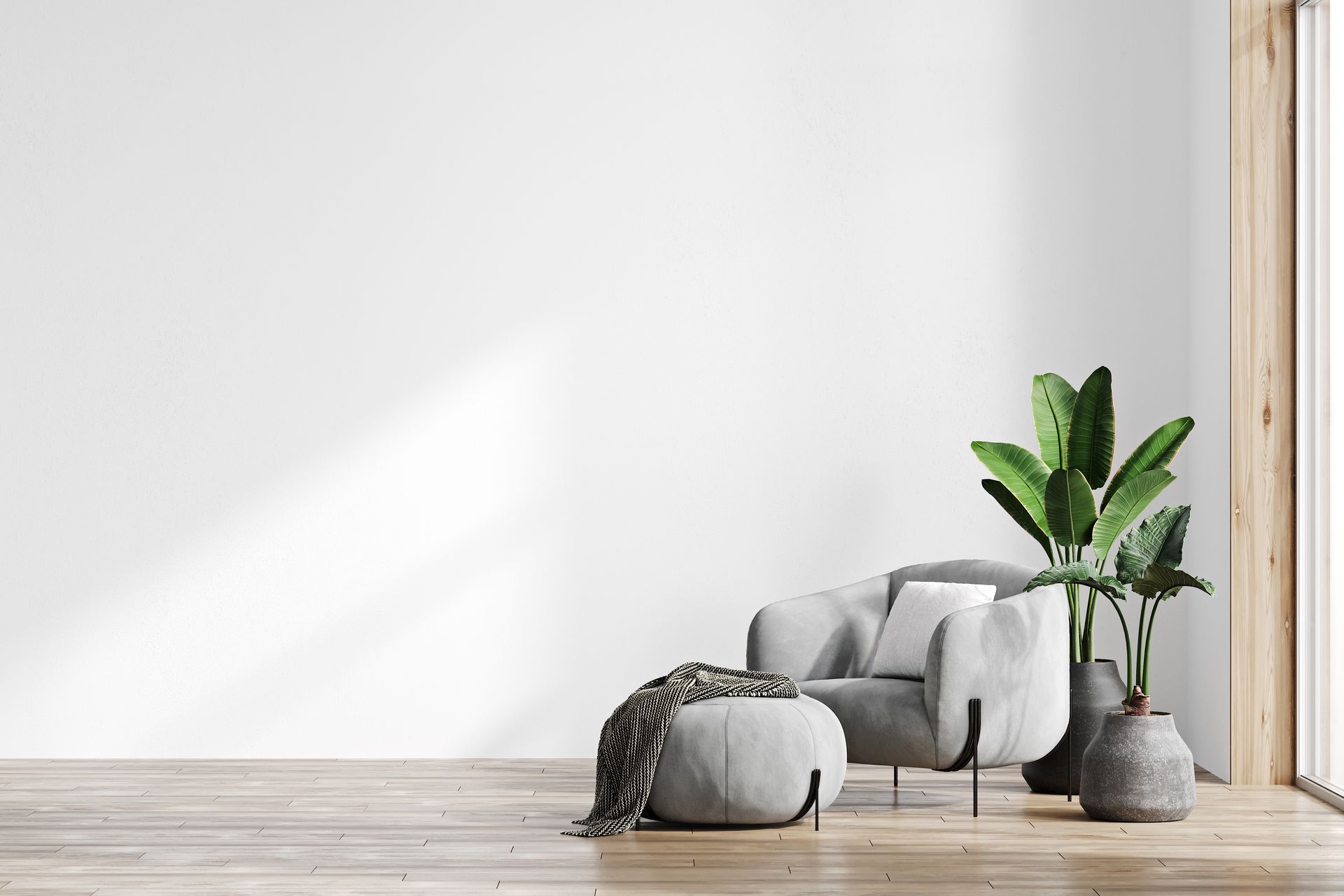 Minimalist living room with a gray chair, pouf, blanket, and potted plant beside a large blank wall.