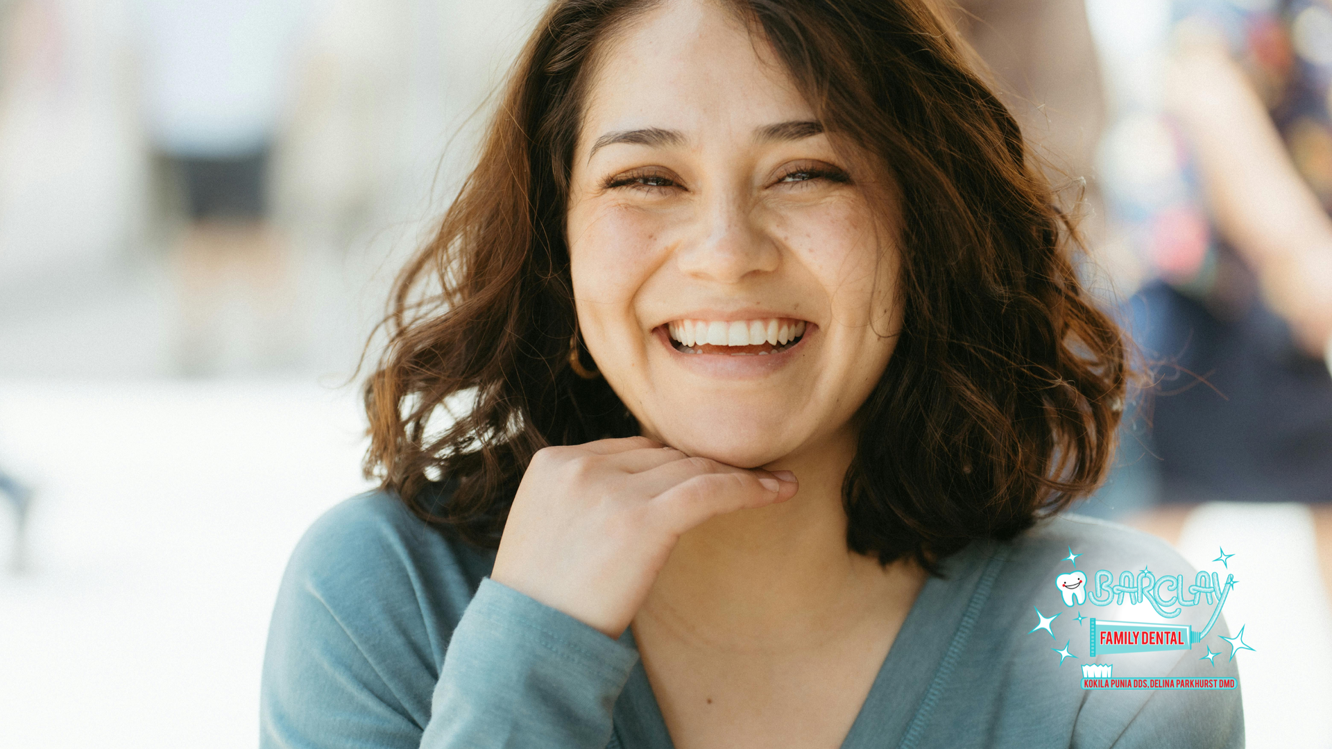 Woman smiling, hand on chin, outdoors, wearing a blue shirt.